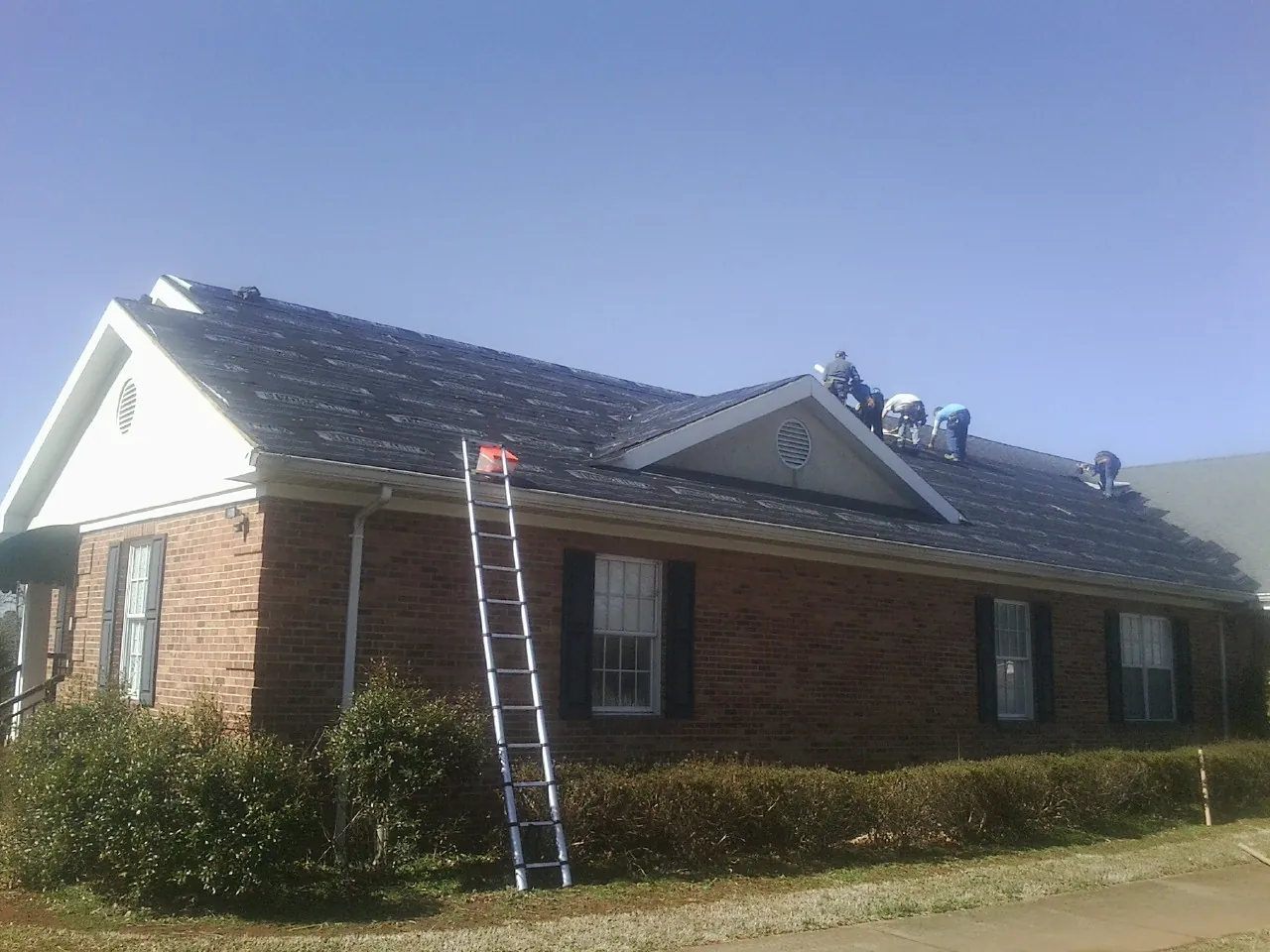 Crew tearing shingles off a brick ranch roof with a ladder set — Emergency Roof Repair work by our crew in Teeds Grove