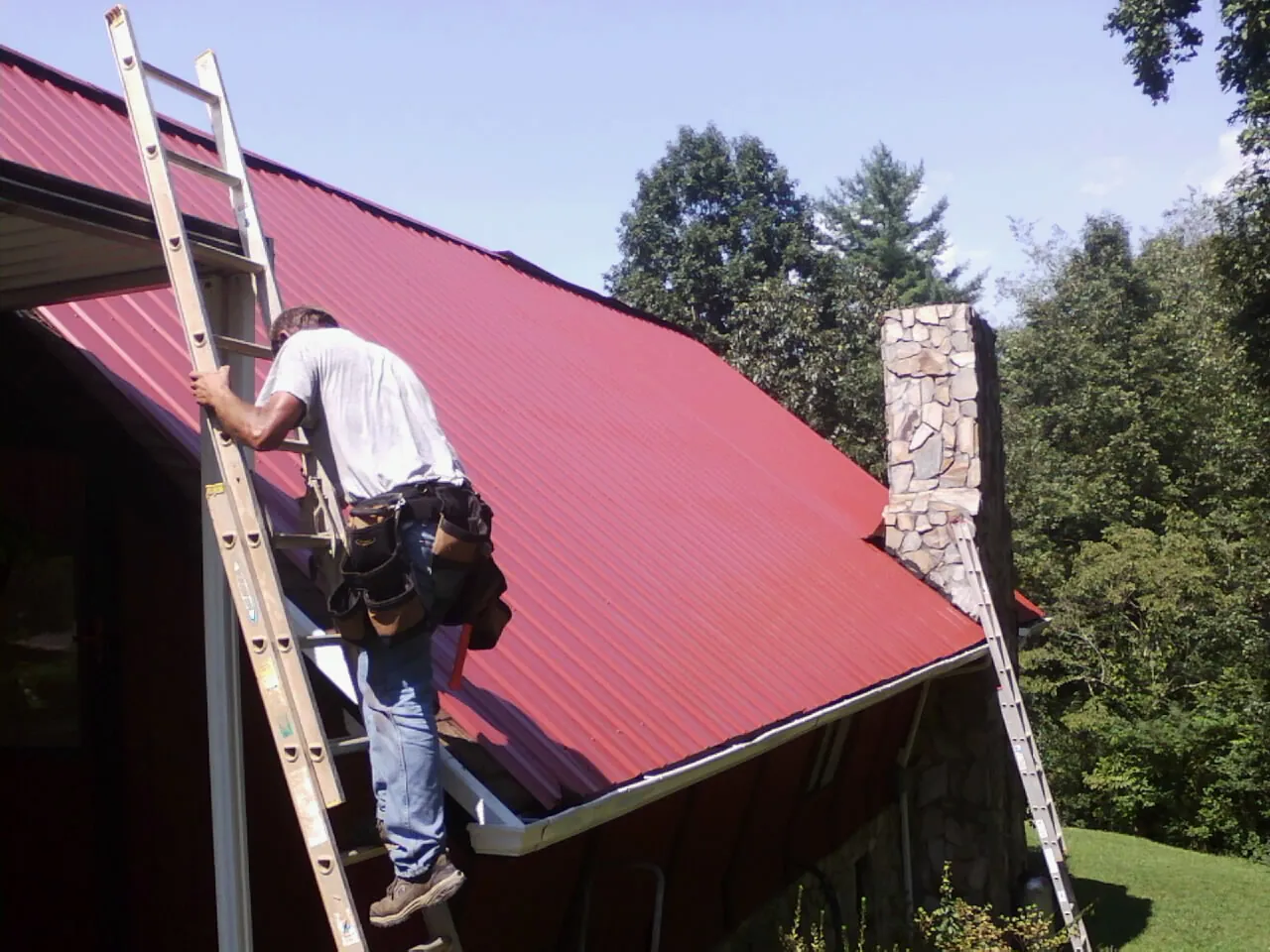 Roofer on a ladder beside a red metal roof and stone chimney — Soffit & Fascia Repair by Roofing Brothers in Teeds Grove